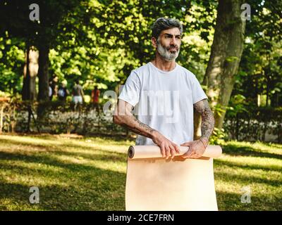 Adult bearded male in activewear rolling up mat after practicing yoga on green lawn in summer park Stock Photo