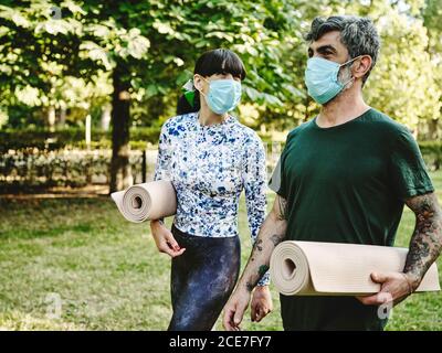 Positive adult tattooed man and cheerful Woman in casual clothes and protective masks with rolled up yoga mats talking while walking in green park after practicing yoga together Stock Photo