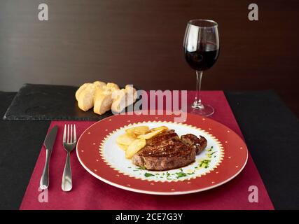Palatable beef steak and potato chips on plate arranged on table with glass of white wine and baguette on slate board in expensive restaurant Stock Photo