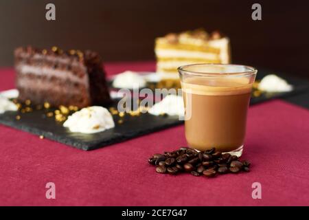 Glass cup of hot latte with coffee beans placed on table near pieces of cakes Stock Photo