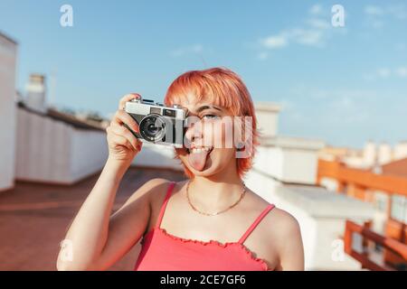 Naughty female millennial with piercing and pink hair taking pictures on vintage photo camera while showing tongue and looking at camera Stock Photo
