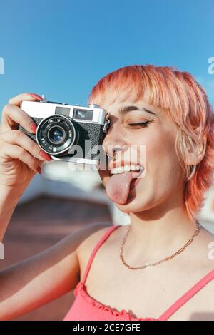 Naughty female millennial with piercing and pink hair taking pictures on vintage photo camera while showing tongue with closed eyes Stock Photo