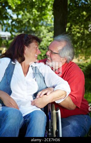 Laughing elderly woman sits in a wheelchair next to her partner in the park Stock Photo