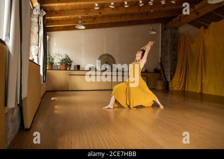 Side view of concentrated female in long waving dressstanding barefoot in studio and practicing yoga in Anjaneyasana Stock Photo