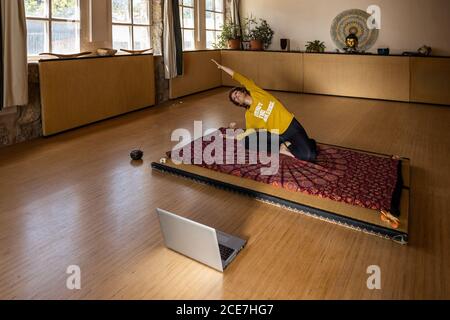 High angle of flexible female sitting on mat in Padmasana and doing side bend while practicing yoga and watching online tutorial via laptop Stock Photo