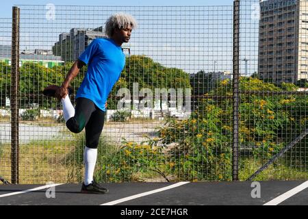 Full length African American sportsman with blond hair standing near net fence and doing quad stretch during fitness workout on sunny day on city street Stock Photo