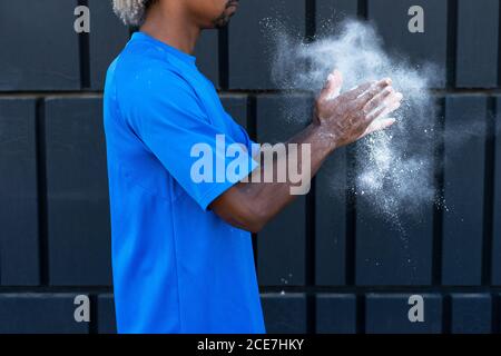 Cropped unrecognizable African American male athlete with dyed hair clapping hands with chalk powder while preparing for training near wall of modern building Stock Photo
