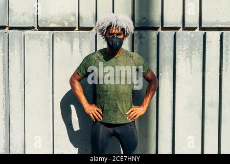 Serious African American male athlete in protective mask holding hands on waist and looking at camera against shabby wall during fitness training on street Stock Photo