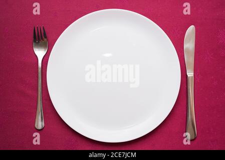 Empty white plate, fork and knife on maroon tablecloth. Elegant table setting. Top view, close-up Stock Photo