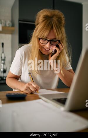 Concentrated female entrepreneur sitting at table in kitchen and working remotely while speaking on cellphone and browsing netbook Stock Photo