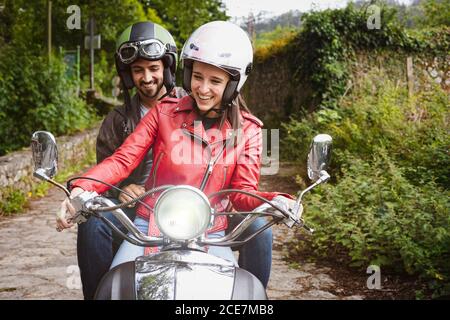 Cheerful man in helmet sitting on motorcycle with smiling girlfriend in leather jacket riding on pathway near lush green shrubs on weekend Stock Photo