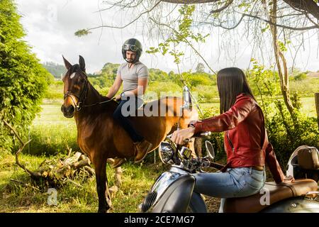 Cheerful man in helmet sitting on horse while interacting with glad girlfriend riding a motorbike near tree in sunlight during trip Stock Photo