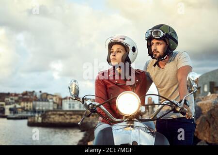 Thoughtful man in helmet standing near wistful girlfriend in leather jacket sitting on motorcycle with headlight on while contemplating picturesque river during vacation and looking away Stock Photo