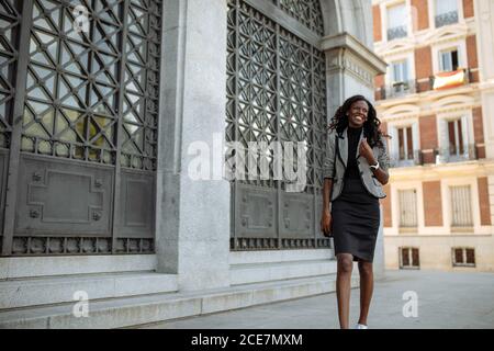 Happy successful young African American business lady in elegant formal outfit walking along old building on city street Stock Photo