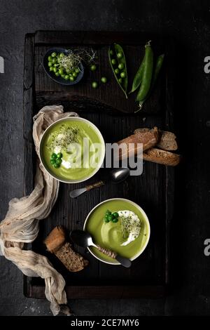 From above view of two bowls of pea cream placed on a wooden tray and dark surface, served with bread and decorated with whole peas and sour cream