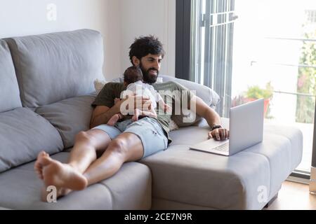 Male freelancer holding baby while lying on sofa and working on project with netbook at home Stock Photo