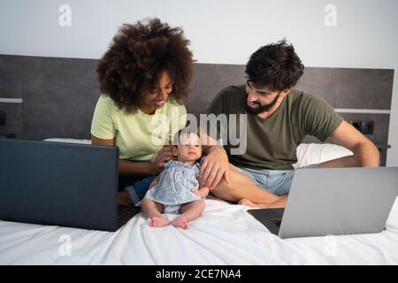 Multiethnic couple of freelancers sitting on bed with cute infant daughter and laptops at home Stock Photo