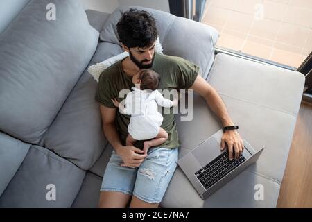 From above male freelancer holding baby while lying on sofa and working on project with netbook at home Stock Photo