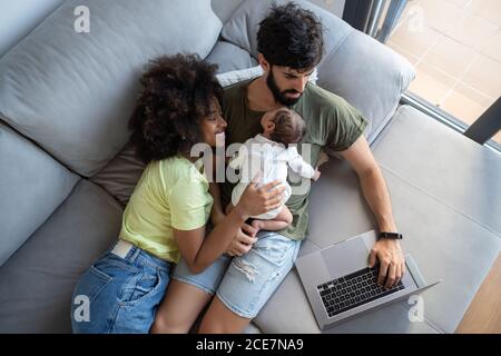 From above of black mother and cute infant lying on couch with Arab father working on laptop at home Stock Photo