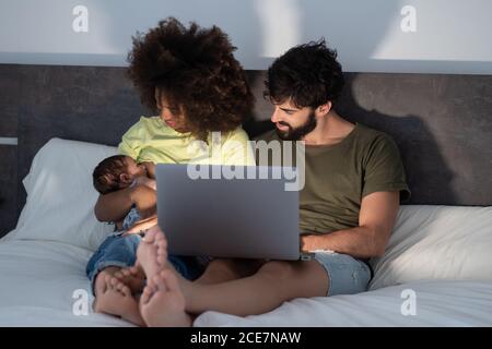 Content Black mother and Arab father sitting on bed with adorable baby and enjoying funny video while using laptop Stock Photo