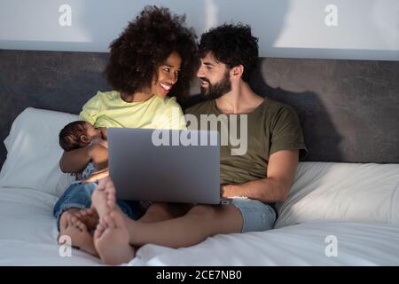 Content Black mother and Arab father sitting on bed with adorable baby and enjoying funny video while using laptop Stock Photo