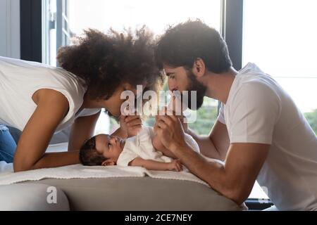 Side view of delighted multiethnic family playing with cute baby on sofa at home