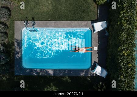 From above drone view of relaxed young female getting inside the water in pool and enjoying summer vacation while spending time in green yard Stock Photo