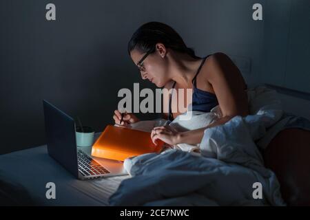 Focused serious female entrepreneur lying on bed and browsing laptop working remotely at night Stock Photo