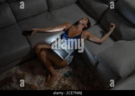 Exhausted female freelancer leaning on couch with laptop and cup of hot drink while working from home remotely Stock Photo