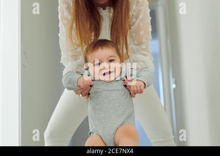 Cute cheerful toddler in gray bodysuit held by crop unrecognizable mother smiling and looking at camera  while having fun together at home Stock Photo