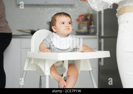 Adorable little baby sitting on high chair and eating fresh avocado from spoon held by crop anonymous mother Stock Photo
