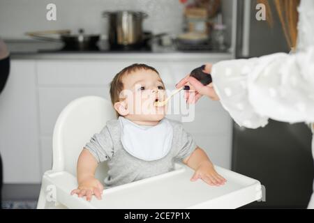 Adorable little baby sitting on high chair and eating fresh avocado from spoon held by crop anonymous mother Stock Photo