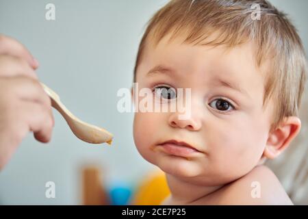 Closeup of adorable little baby eating puree from spoon held by crop anonymous mother Stock Photo