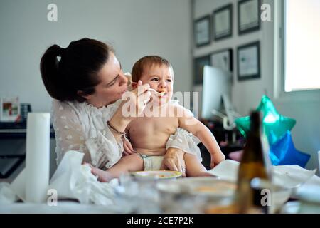 Crying little baby sitting on knees of mother and refusing food from spoon during feeding time at home Stock Photo