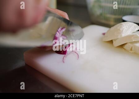 From above of crop anonymous chef cutting red onion in half circles using knife and cutting board near fresh mozzarella cheese while cooking Stock Photo