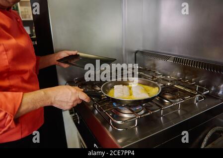 Crop unrecognizable chef passing fish fillets in melted butter and olive oil in frying pan using fork and spoon while preparing food at home Stock Photo