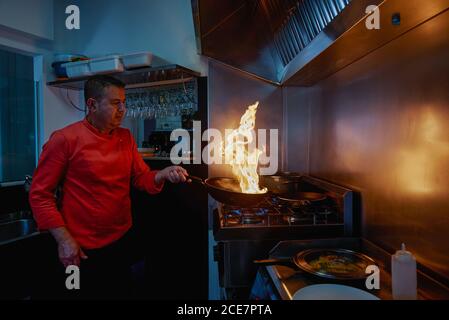 Concentrated chef in uniform holding frying pan while flambeing ingredients in frying pan on slow heat standing in restaurant kitchen in evening and looking at bright fire Stock Photo