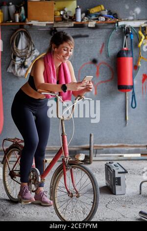 Full body smiling female bicyclist in sportswear sitting on bicycle and browsing mobile phone in messy garage while resting after ride Stock Photo