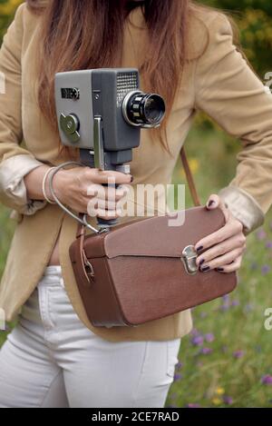 Crop unrecognizable slim female photographer in jacket standing on grass with leather case and retro video camera in countryside in summer Stock Photo