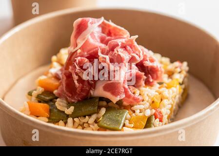 Closeup of tasty rice with various vegetables garnished with Iberico ham and placed in cardboard bowl on table Stock Photo
