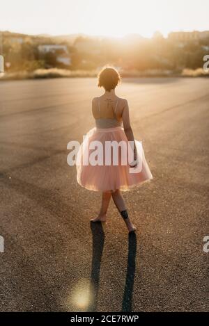 Full body of delighted unrecognizable female artist in elegant dance dress laughing and dancing in back lit in empty square with blurred town in background in sunny summer sunset day Stock Photo