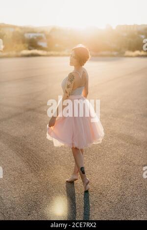 Full body delighted young female artist in elegant dance dress laughing and dancing in back lit in empty square with blurred town in background in sunny summer sunset day Stock Photo