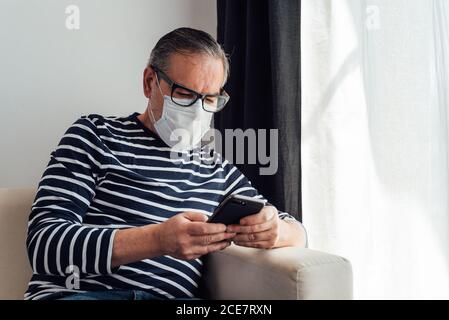 Focused male in glasses using on mobile phone and laptop while working at home because of quarantine Stock Photo