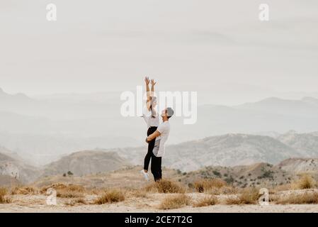 Side view of happy young man holding excited girlfriend while standing on dry hill against foggy mountains during romantic journey together in nature Stock Photo