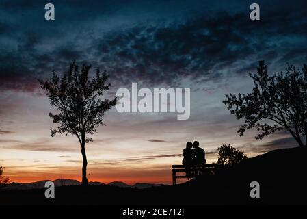 Back view silhouettes of affectionate couple enjoying romantic date while sitting on bench against cloudy sunset sky in summer evening Stock Photo