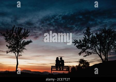Back view silhouettes of affectionate couple enjoying romantic date while sitting on bench against cloudy sunset sky in summer evening Stock Photo