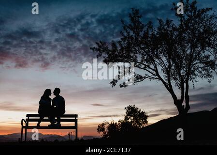 Back view silhouettes of affectionate couple enjoying romantic date while sitting on bench against cloudy sunset sky in summer evening Stock Photo