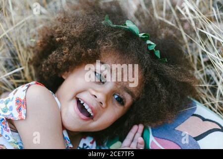 Adorable calm mulatto girl in trendy dress lying on plaid on wheat field on summer day and looking at camera Stock Photo