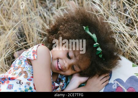 Adorable calm mulatto girl in trendy dress lying on plaid on wheat field on summer day and looking at camera Stock Photo