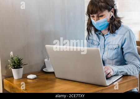 Focused female freelancer in denim shirt and medical mask using earbuds and working remotely in cafe using laptop during coronavirus epidemic Stock Photo
